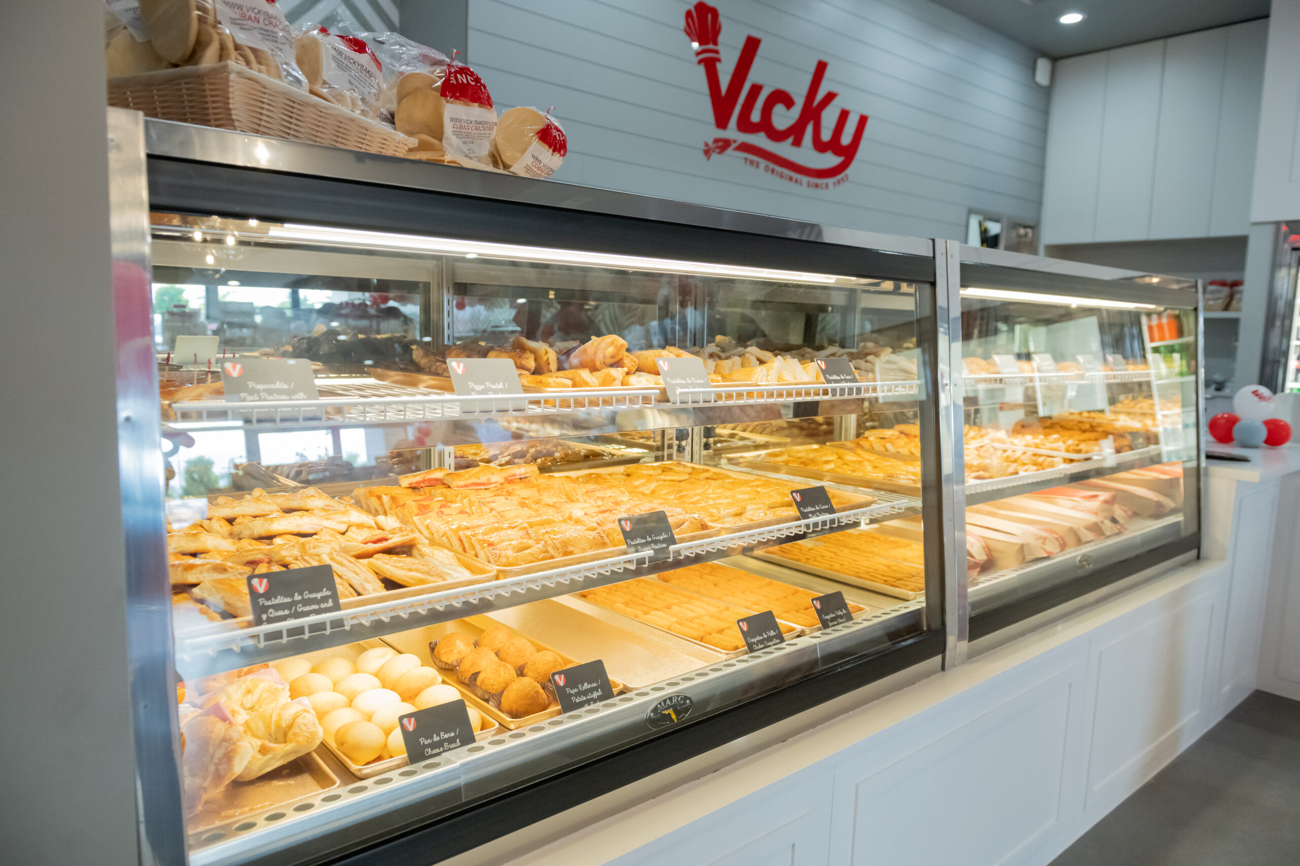 Two people seated at a marble table enjoying Vicky Bakery items, including pastelitos in branded red and white boxes, a hot coffee in a red cup, and an iced coffee